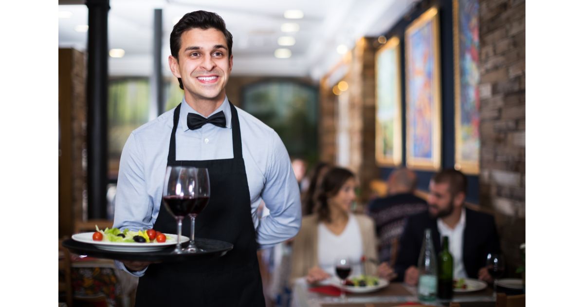 A smiling waiter in a black apron and bow tie serving a plate of salad and a glass of red wine at a restaurant, with diners enjoying their meals in the background.