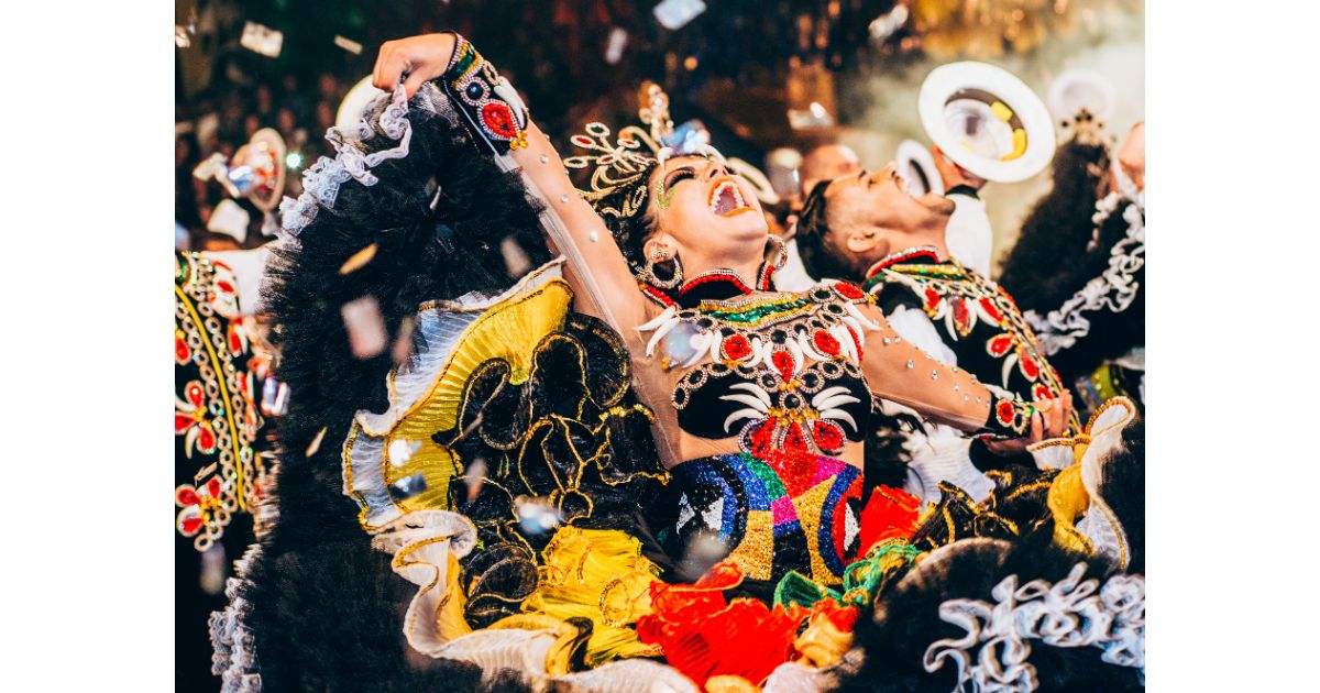 Two performers in vibrant, colorful costumes dancing joyfully, surrounded by confetti, at a lively festival or carnival celebration.