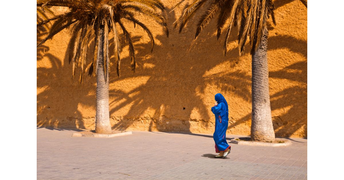 A woman dressed in a blue traditional garment walking past palm trees against a golden wall, casting long shadows on the ground.