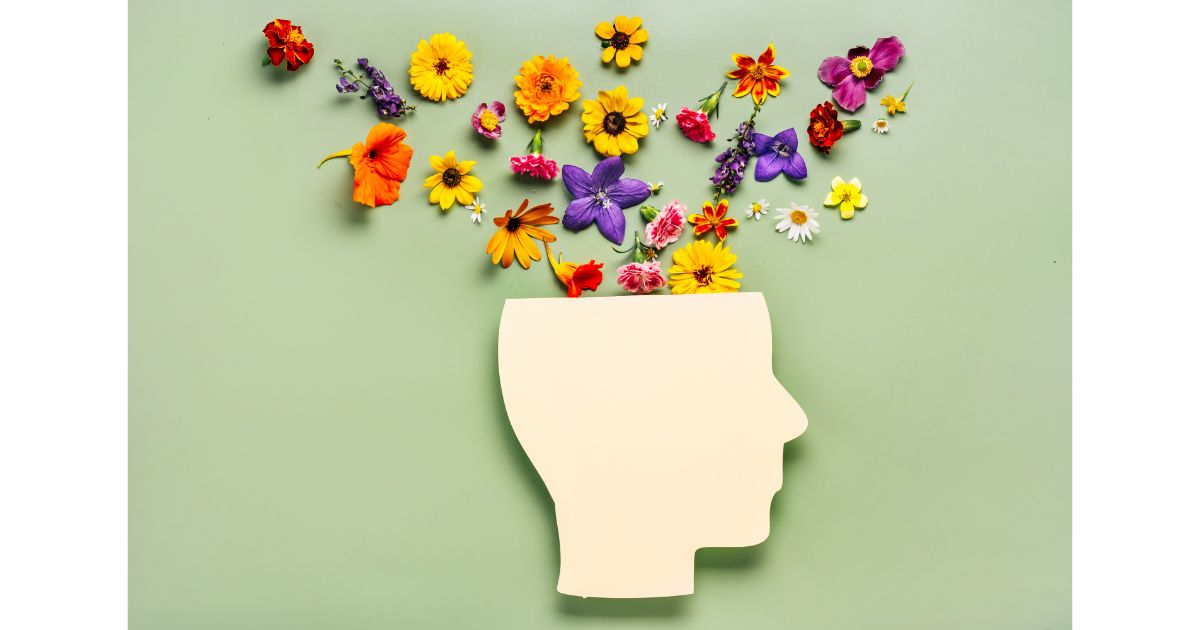 A silhouette of a human head with colorful flowers blooming from the top, symbolizing creativity, growth, and the flourishing of ideas. The background is a soft green.