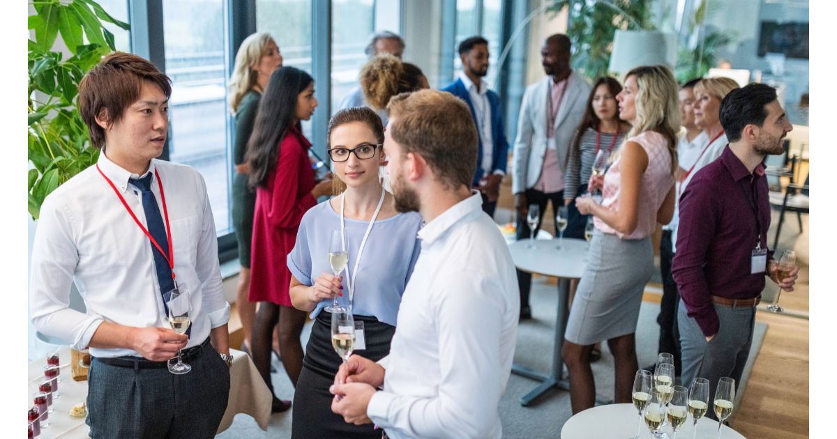 A group of people in business attire engaging in conversation at a corporate event. Some attendees are holding glasses of champagne, while others are mingling and networking in a modern office setting.