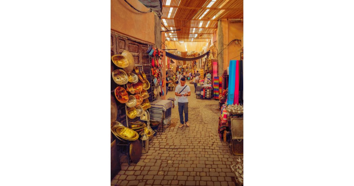 A vibrant market street in Marrakesh with colorful goods, including hats and textiles, and a person walking through the cobbled path.