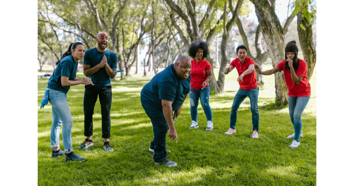 A group of six diverse adults playing a fun outdoor team game in a park, wearing red and blue shirts, laughing and cheering under the shade of tall trees.