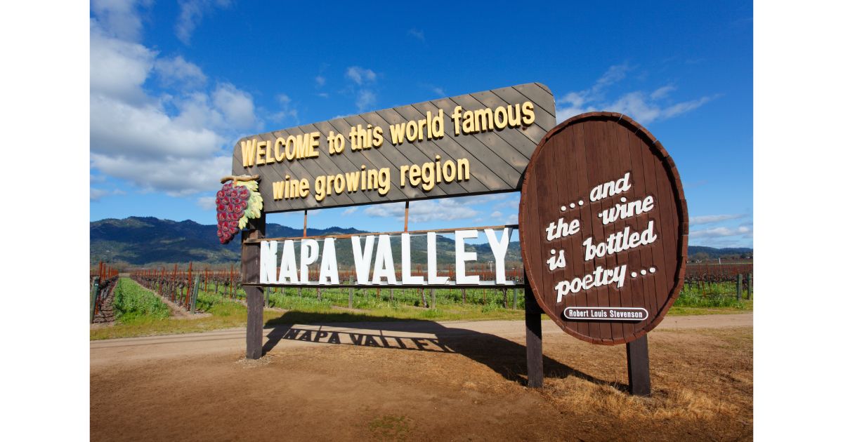 Iconic Napa Valley welcome sign with a wooden plaque that reads 'Welcome to this world-famous wine-growing region' and a quote by Robert Louis Stevenson, set against a scenic vineyard and blue sky.