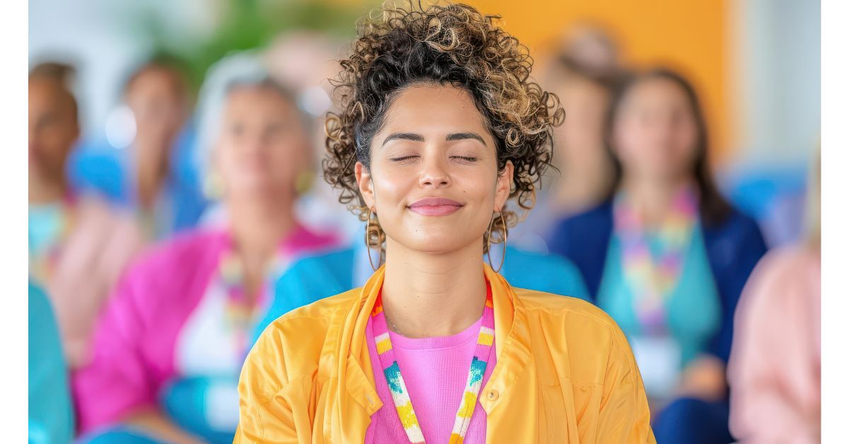 A young woman with curly hair, wearing a colorful outfit, sits with her eyes closed and a peaceful smile, surrounded by a blurred group of people in a vibrant setting.