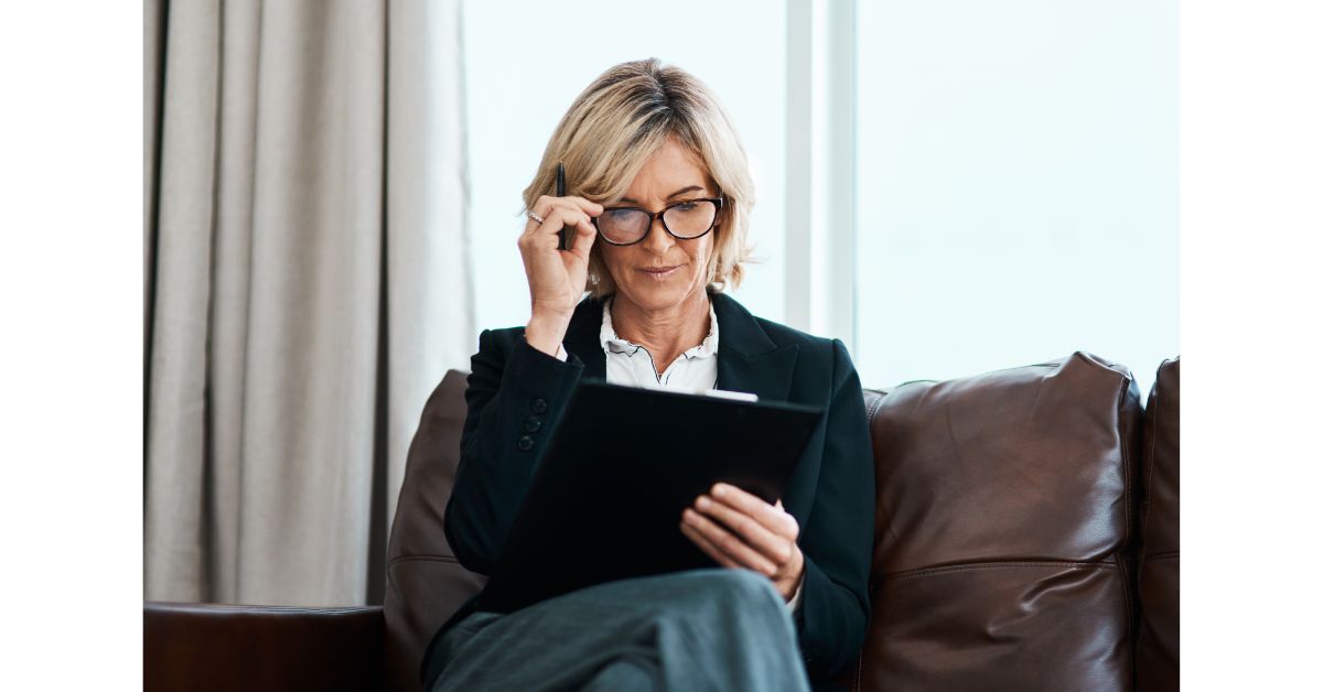 A professional woman with blonde hair and glasses, dressed in business attire, sits on a leather couch, reviewing documents on a clipboard with a focused expression.