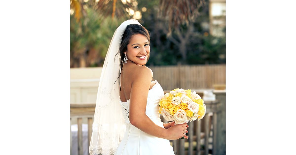 A smiling bride holding a bouquet of yellow and white roses, looking over her shoulder while wearing a wedding dress and veil.