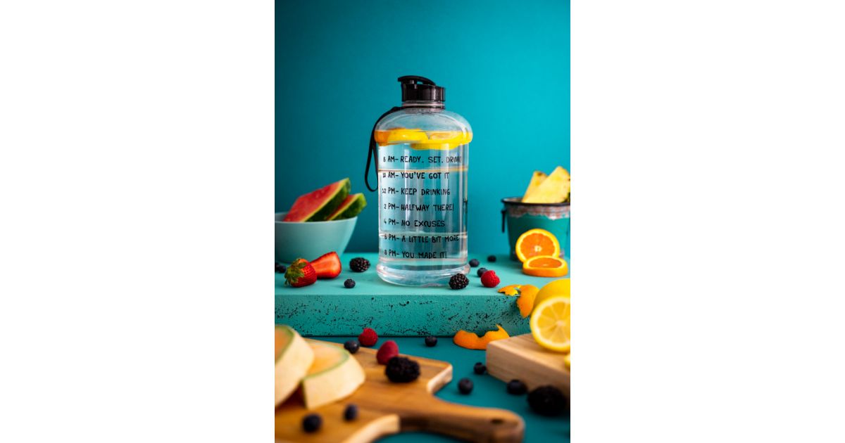 A large water bottle with time markers for hydration goals, surrounded by fresh fruit including watermelon, strawberries, oranges, and berries, set against a vibrant blue background.