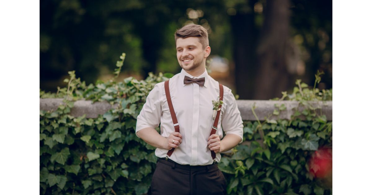 A groom smiling while adjusting his suspenders and bow tie, standing outdoors with greenery in the background, wearing a boutonniere on his shirt.