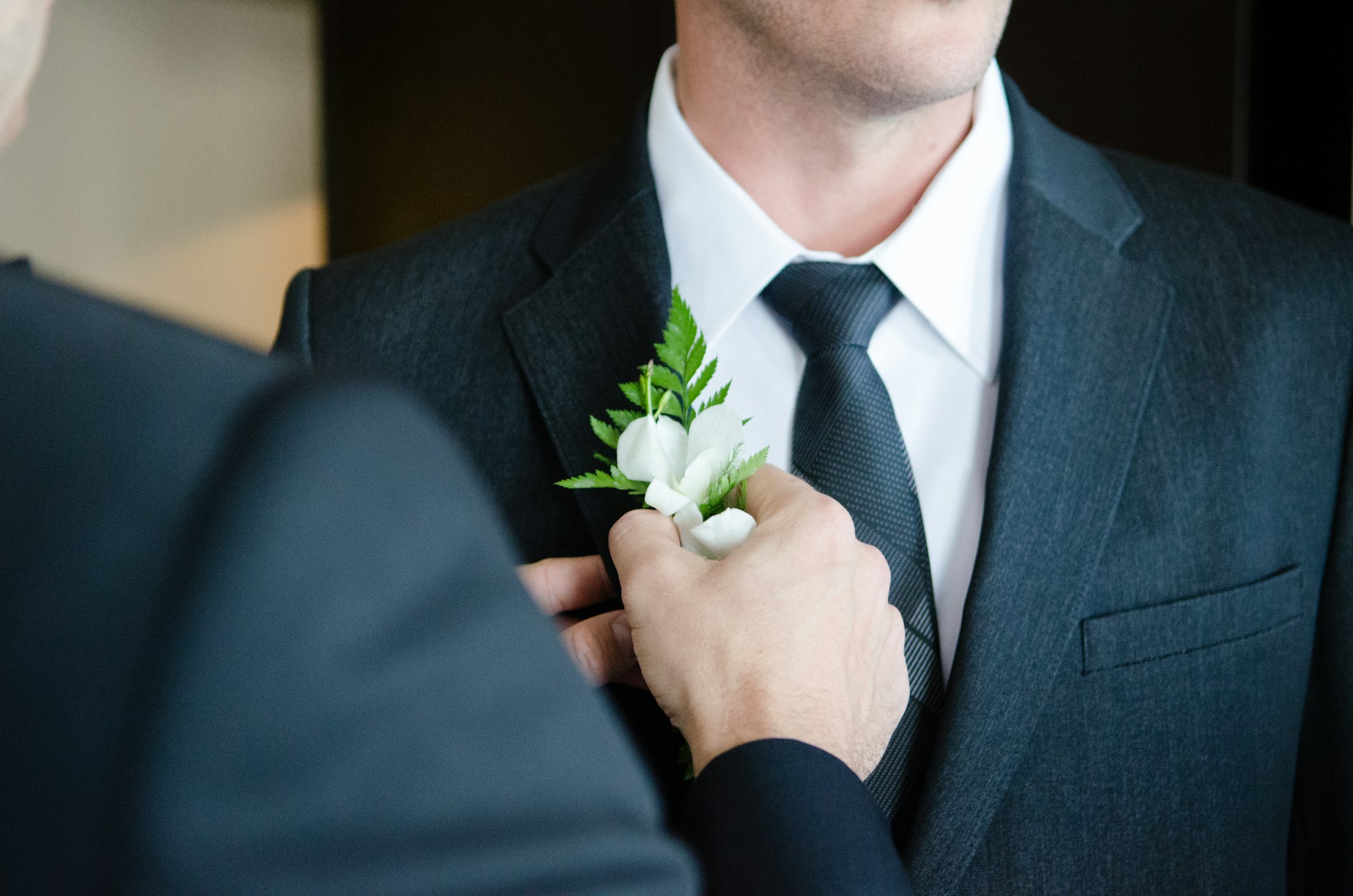 A close-up of a groom having a boutonniere attached to his suit by another person, showcasing a moment of preparation before the wedding ceremony.