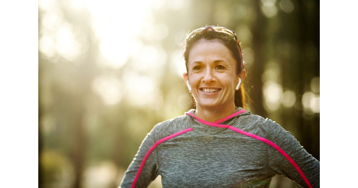 A woman smiling while standing outdoors, wearing a workout top and wireless earbuds.