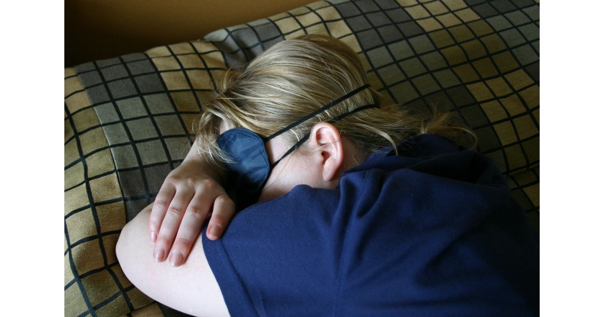 A person wearing an eye mask, resting on a bed with their arms crossed.