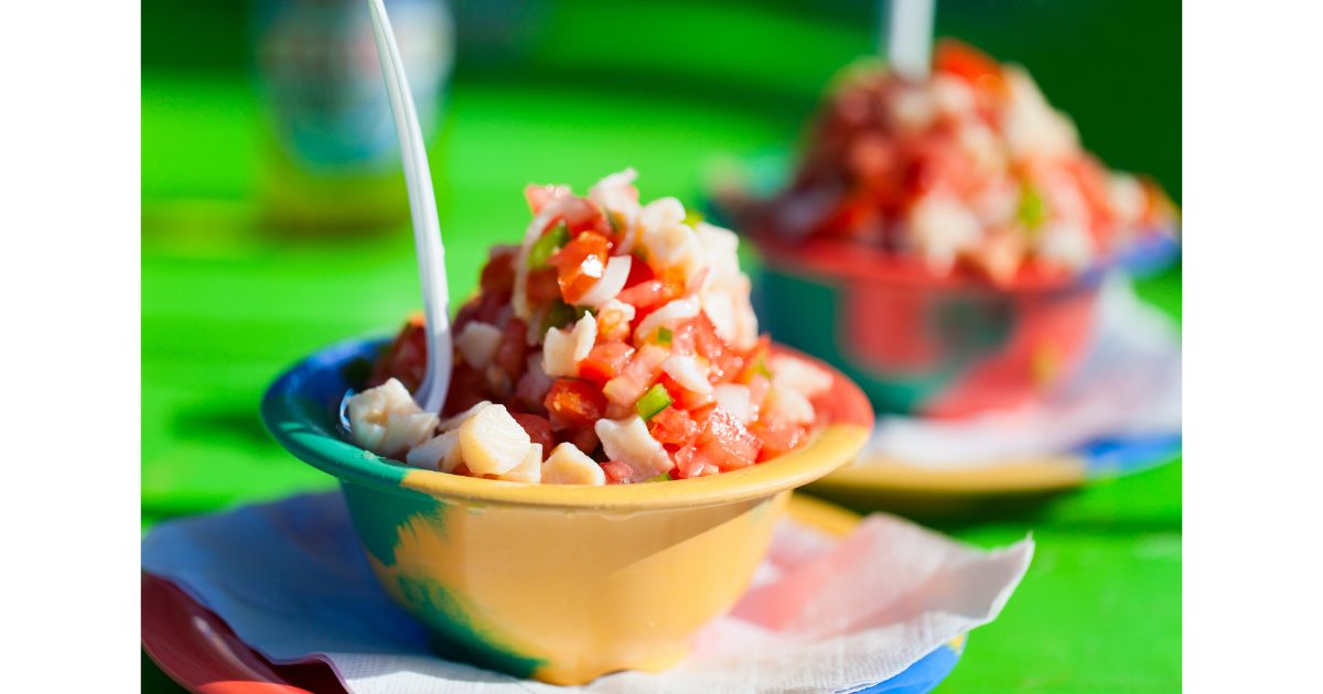 Close-up of a colorful bowl of fresh conch salad with diced tomatoes, onions, and peppers, served with a spoon on a vibrant green table.