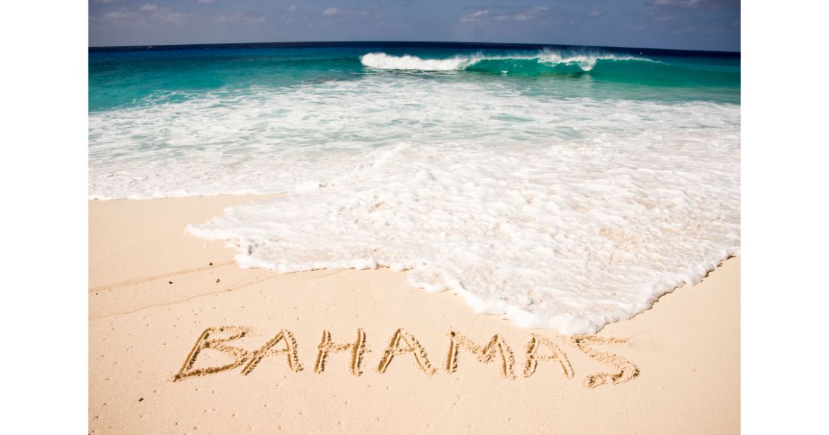 Beach scene with the word 'Bahamas' written in the sand as ocean waves approach, under a bright blue sky.