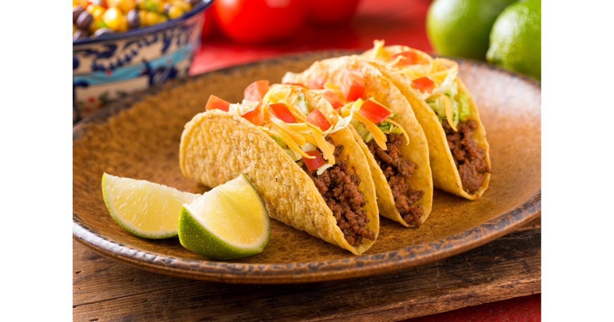 Three delicious tacos filled with seasoned ground beef, topped with shredded lettuce, diced tomatoes, and cheese, served with lime wedges on the side.