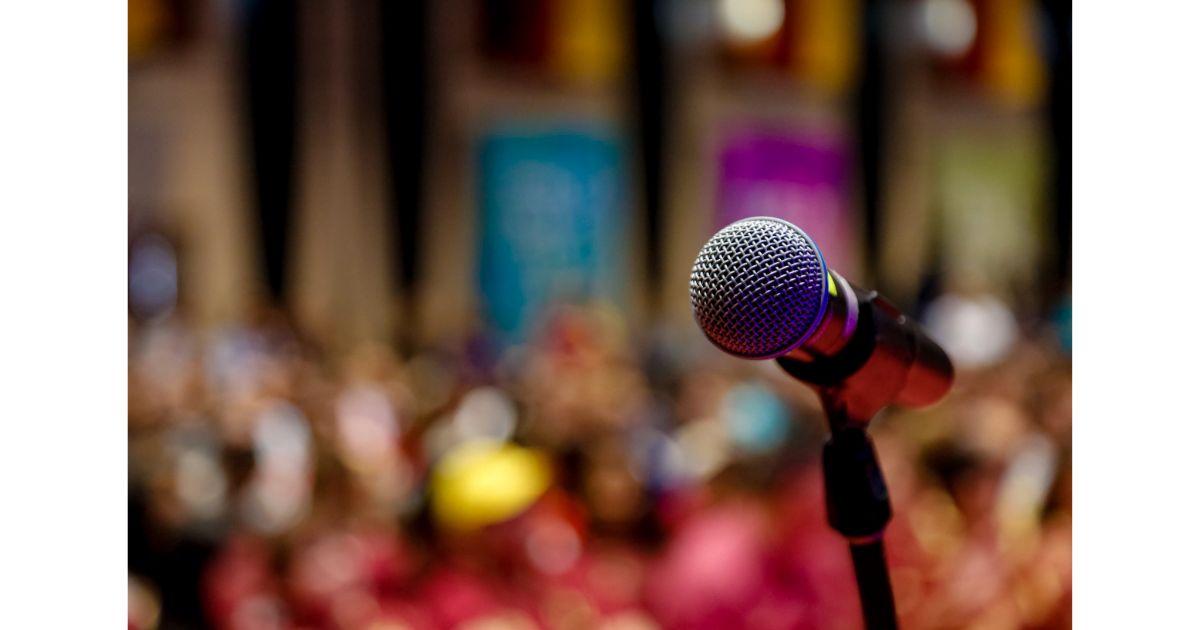 A close-up of a microphone in focus with a blurred crowd in the background, capturing the anticipation of a live event or performance.