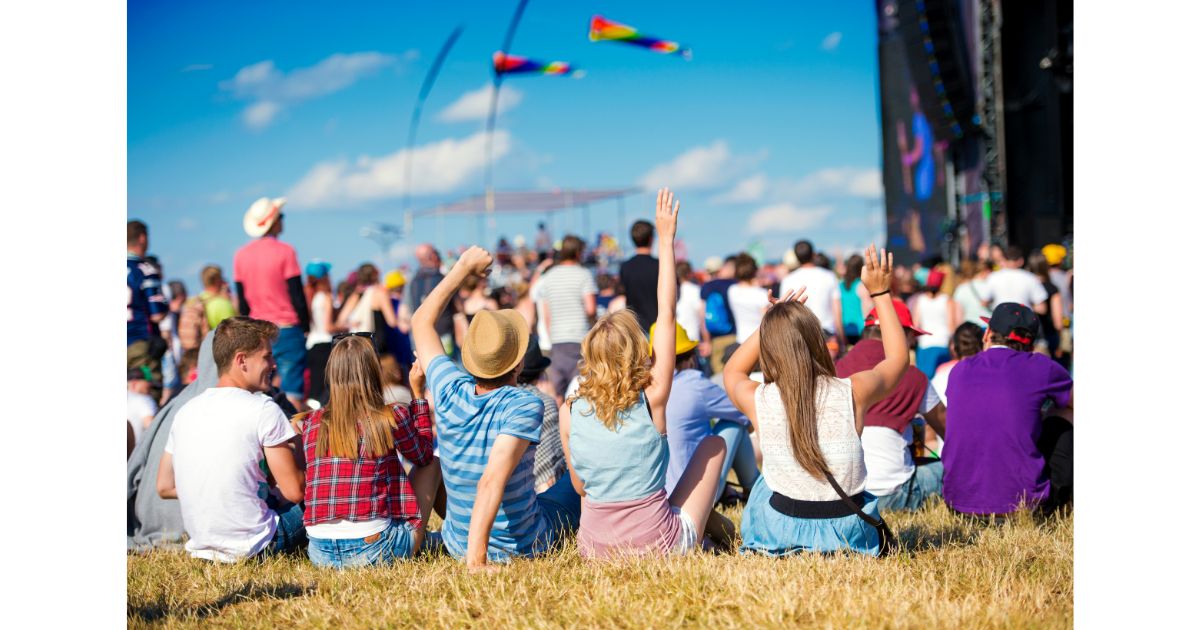 A group of festival-goers sitting on the grass with their arms raised, enjoying a live performance at an outdoor music festival on a sunny day.