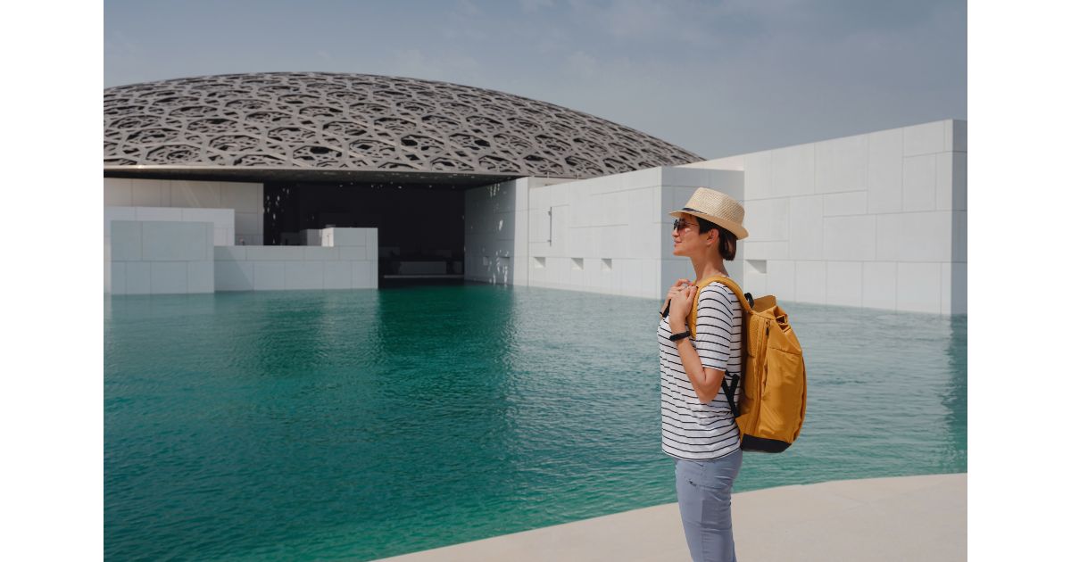 A woman with a yellow backpack and a straw hat standing near the water at the Louvre Abu Dhabi, with the iconic dome structure in the background.