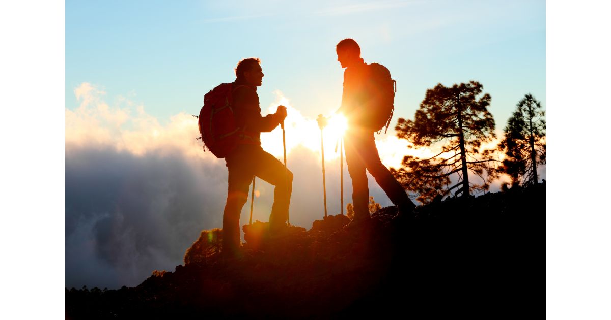 Two hikers standing on a mountain trail at sunset, with backpacks and trekking poles, silhouetted against the beautiful sky and trees.