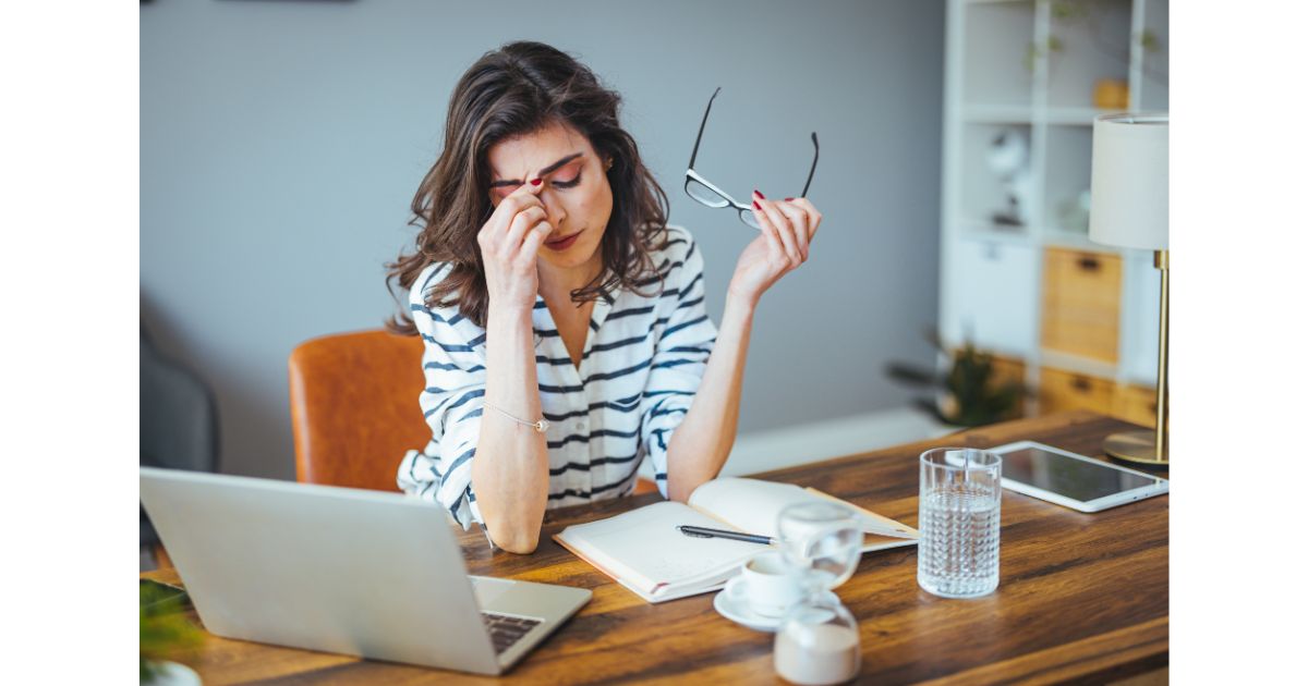 Stressed woman at work holding her head while removing glasses, sitting at a desk with a laptop, notebook, and glass of water, showing signs of fatigue.