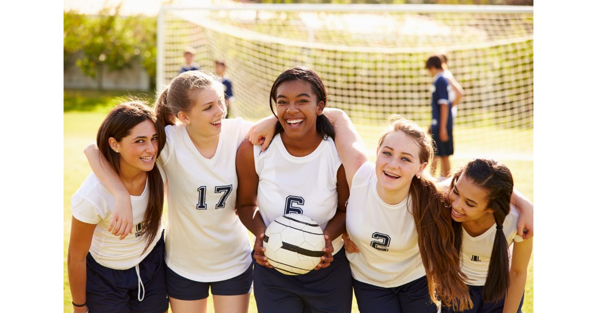 A group of five young female soccer players smiling and posing together on the field, holding a soccer ball and wearing matching team jerseys.