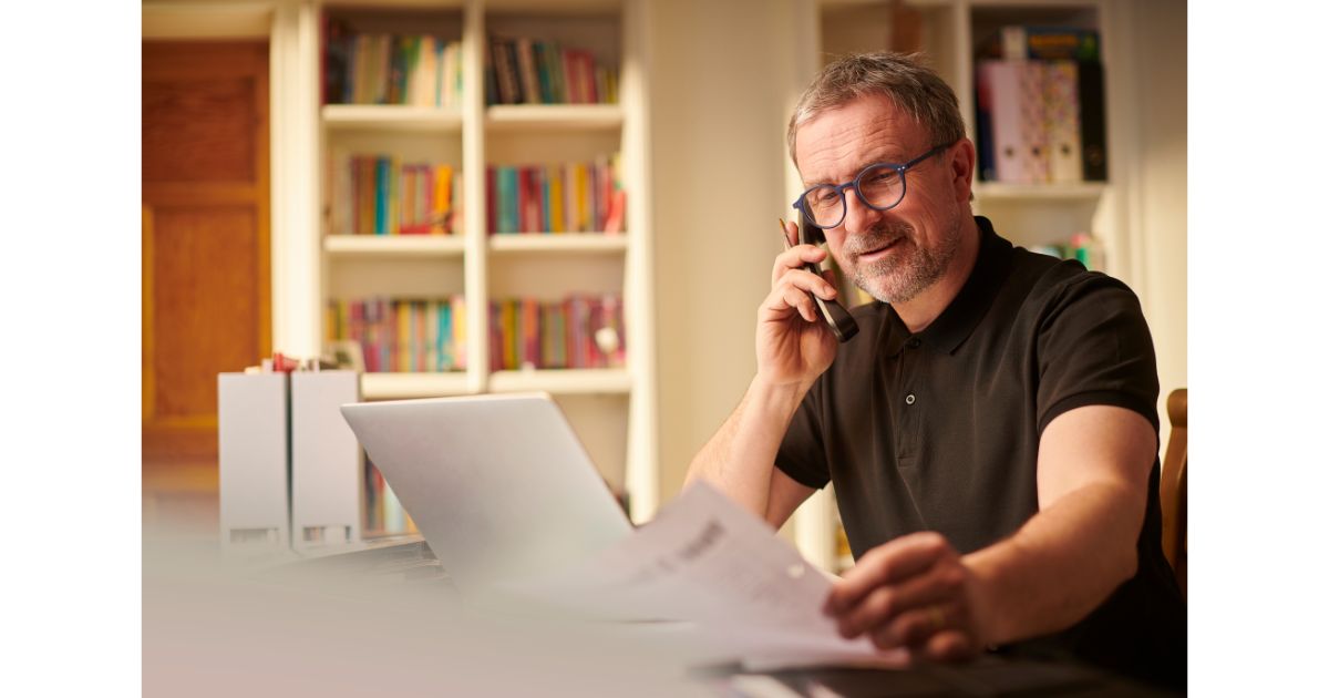 A man sitting at his desk, talking on the phone while reviewing papers, with a laptop open in front of him and bookshelves in the background.