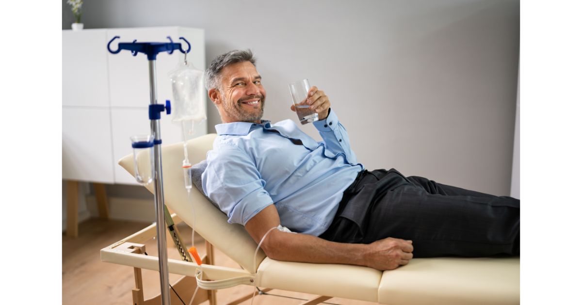 A man receiving IV hydration therapy while relaxing on a treatment table, holding a glass of water and smiling.