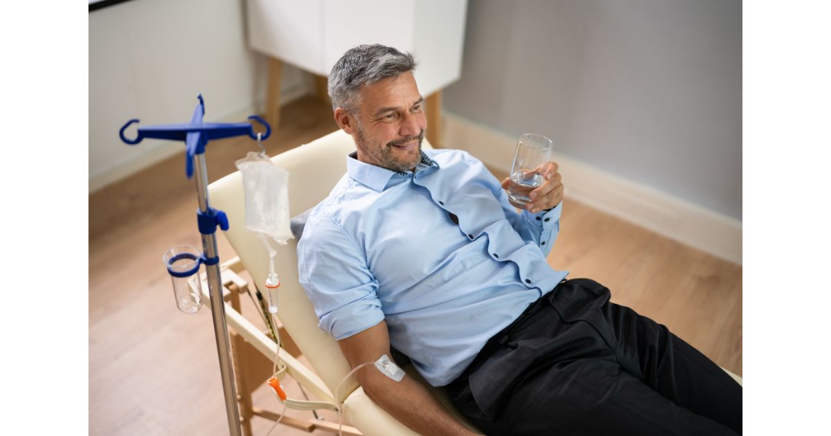 A man relaxing in a chair while receiving an IV drip, holding a glass of water, and enjoying the treatment in a calm environment.