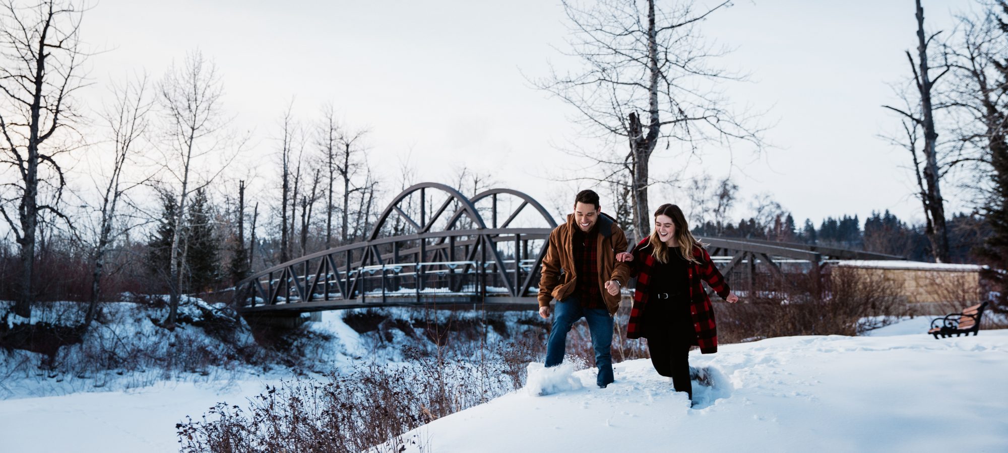 A couple walking hand-in-hand across a bridge in a snowy landscape, enjoying a winter day in Edmonton, with bare trees surrounding them.