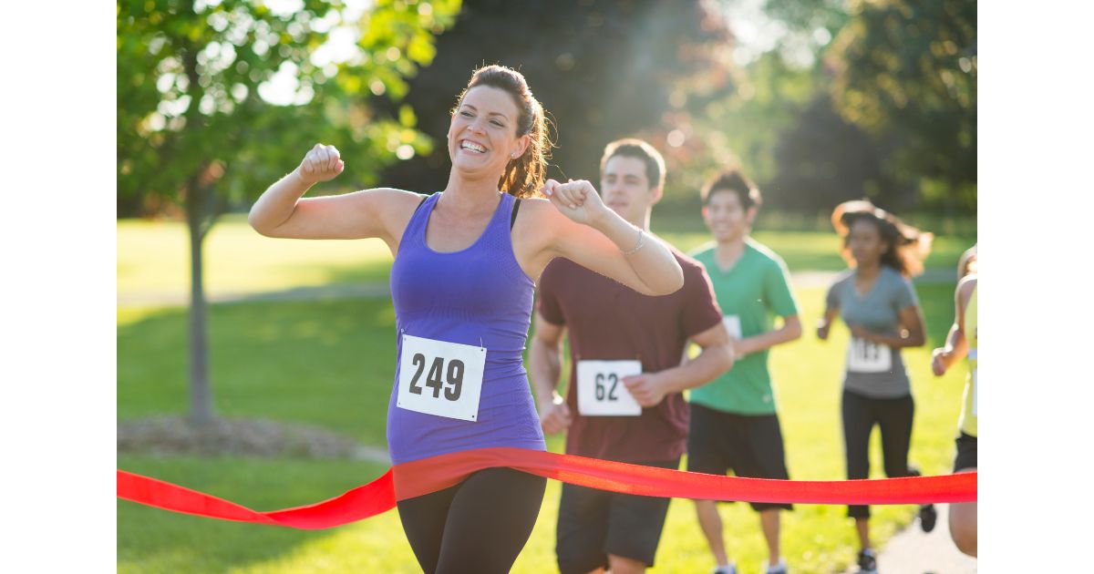 A woman crossing the finish line with a big smile at a marathon, surrounded by other participants in a sunny outdoor setting.