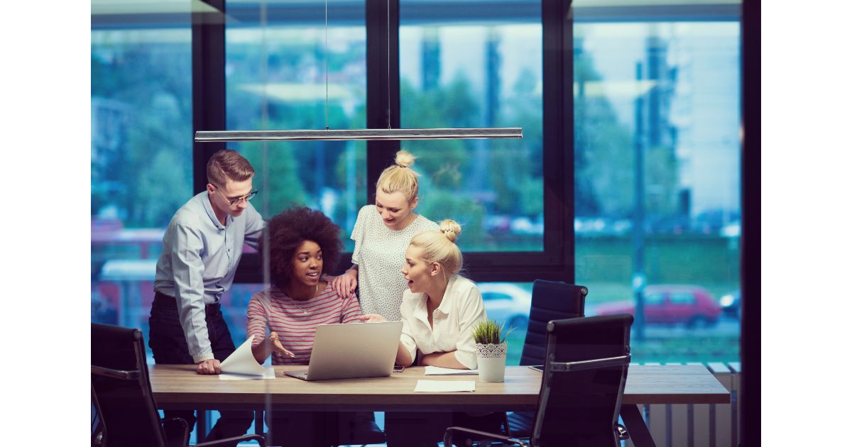 A diverse group of four colleagues collaborating around a laptop in a modern office, discussing ideas and working together in a professional setting.