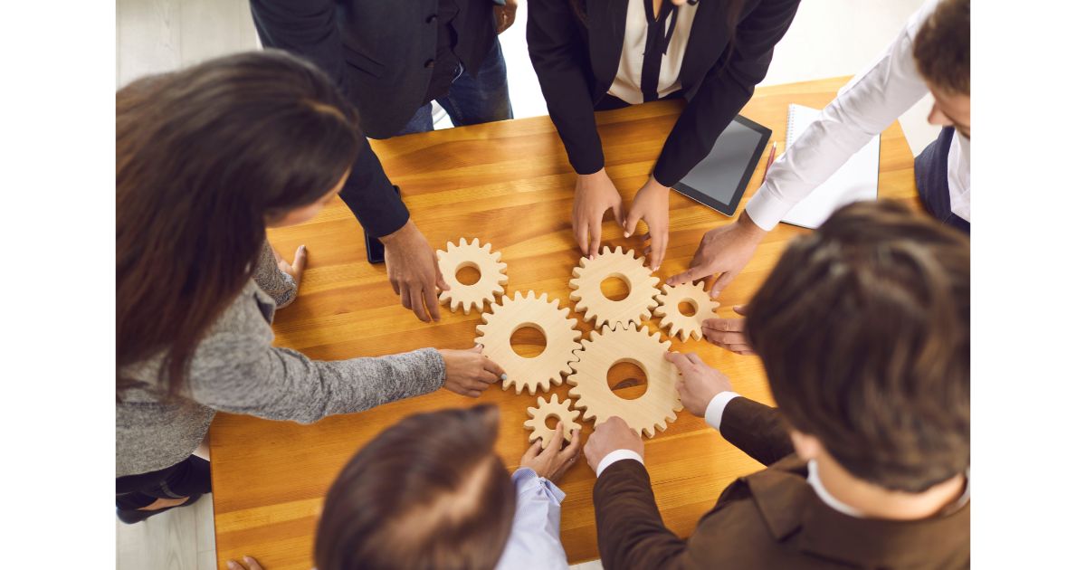 A group of professionals working together, placing wooden gear pieces on a table to symbolize collaboration and teamwork in business or problem-solving.