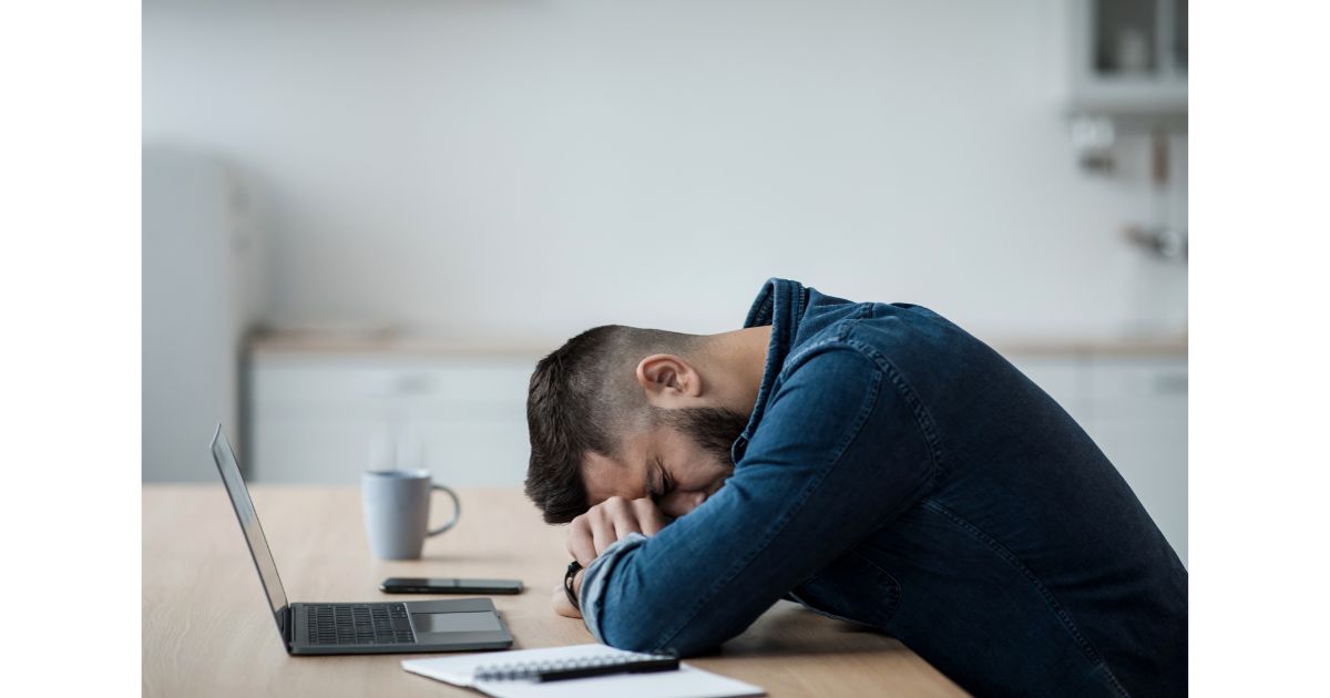 A man sitting at a desk, resting his head on his arms, appearing stressed or exhausted while surrounded by a laptop, phone, and notepad.