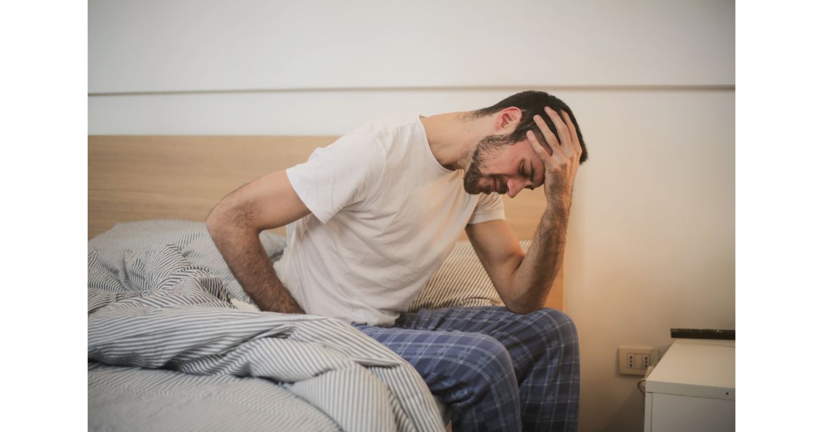 A man sitting on the edge of a bed, holding his head in discomfort, possibly experiencing a headache or hangover symptoms.