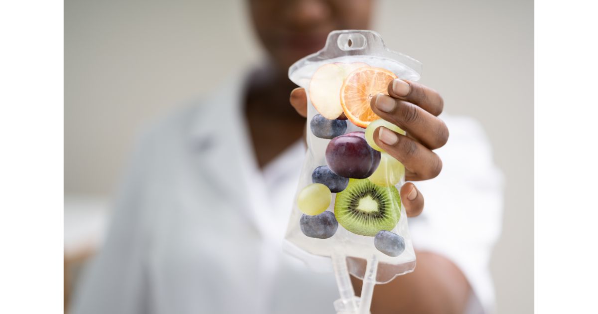 A close-up of a person holding an IV bag filled with slices of fruit, including kiwi, grapes, and citrus, symbolizing health and wellness.