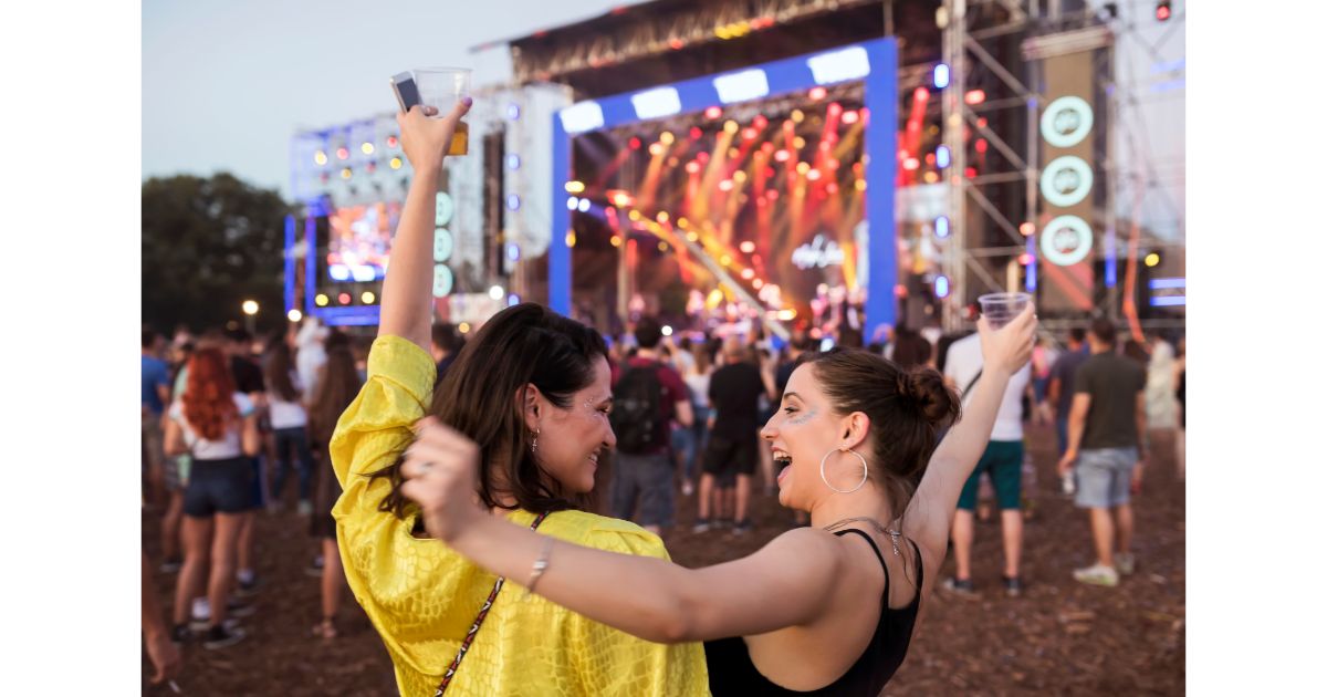 Two women enjoying a music festival, raising their arms and holding drinks, with a vibrant stage and crowd in the background.