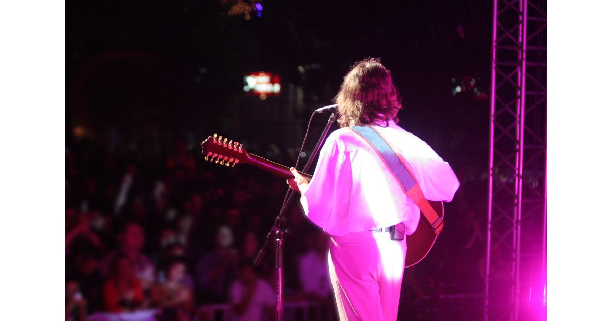 A musician performing on stage with a guitar, facing away from the audience under vibrant stage lights. The crowd can be seen in the background, enjoying the performance.