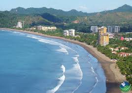 A scenic view of Jacó Beach, showing the coastline with gentle waves, sandy shores, and nearby buildings, set against a backdrop of lush green mountains.