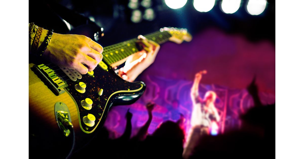 A close-up of a guitarist playing on stage with vibrant lighting in the background, while a blurred performer can be seen in the distance, capturing the energy of a live music performance.