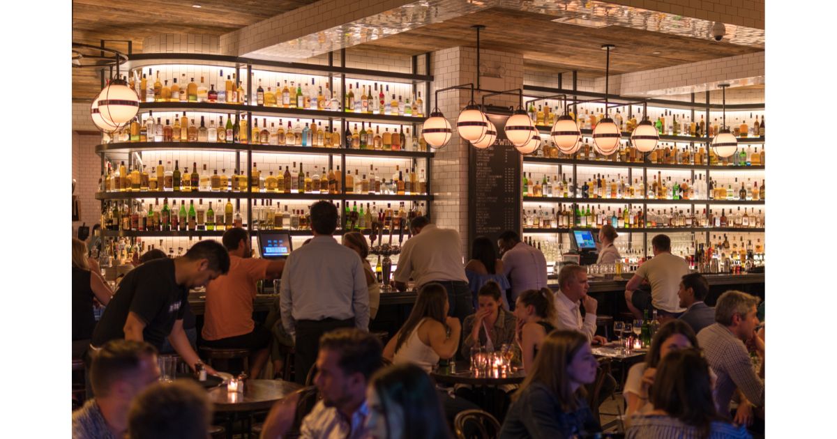 A bustling bar scene with shelves lined with various bottles of alcohol, a bartender serving drinks, and people socializing at tables in a warm, ambient setting.