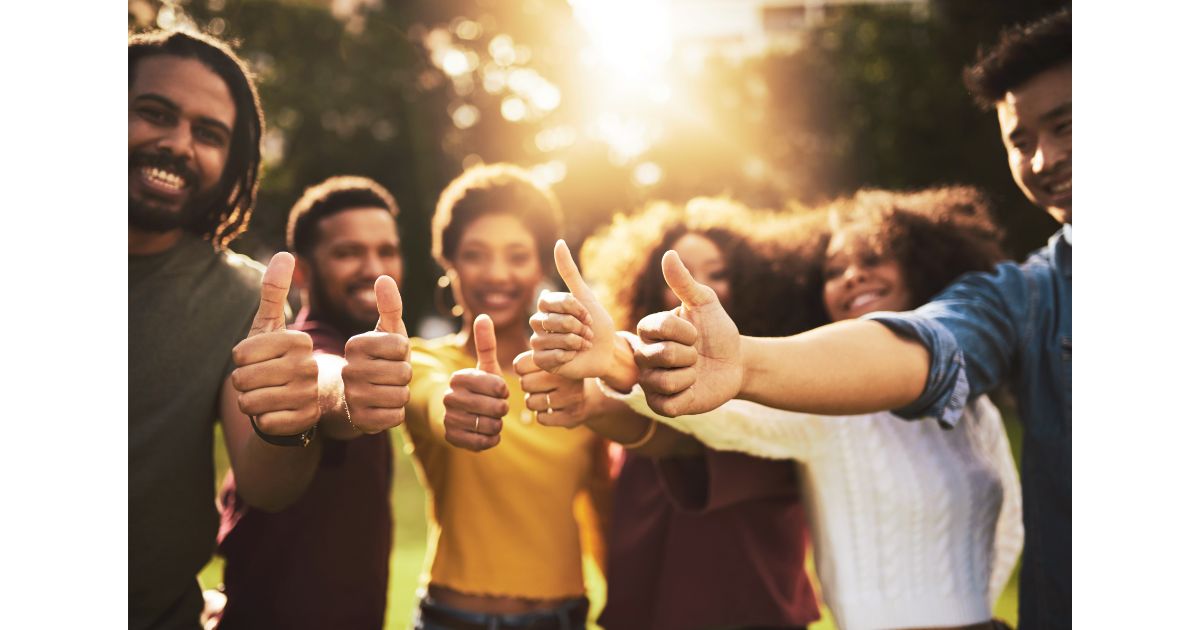 A group of six people standing together in a sunny outdoor setting, all giving a thumbs-up with smiles on their faces, showcasing a positive and friendly atmosphere.