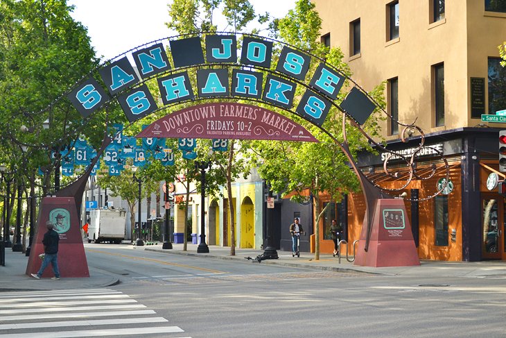 A vibrant entrance arch for the San Jose Sharks, located near the Downtown Farmers Market, with trees lining the street and nearby shops.