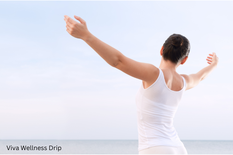 Woman in white tank top stretching arms out toward the ocean, viewed from behind, symbolizing freedom and wellness.