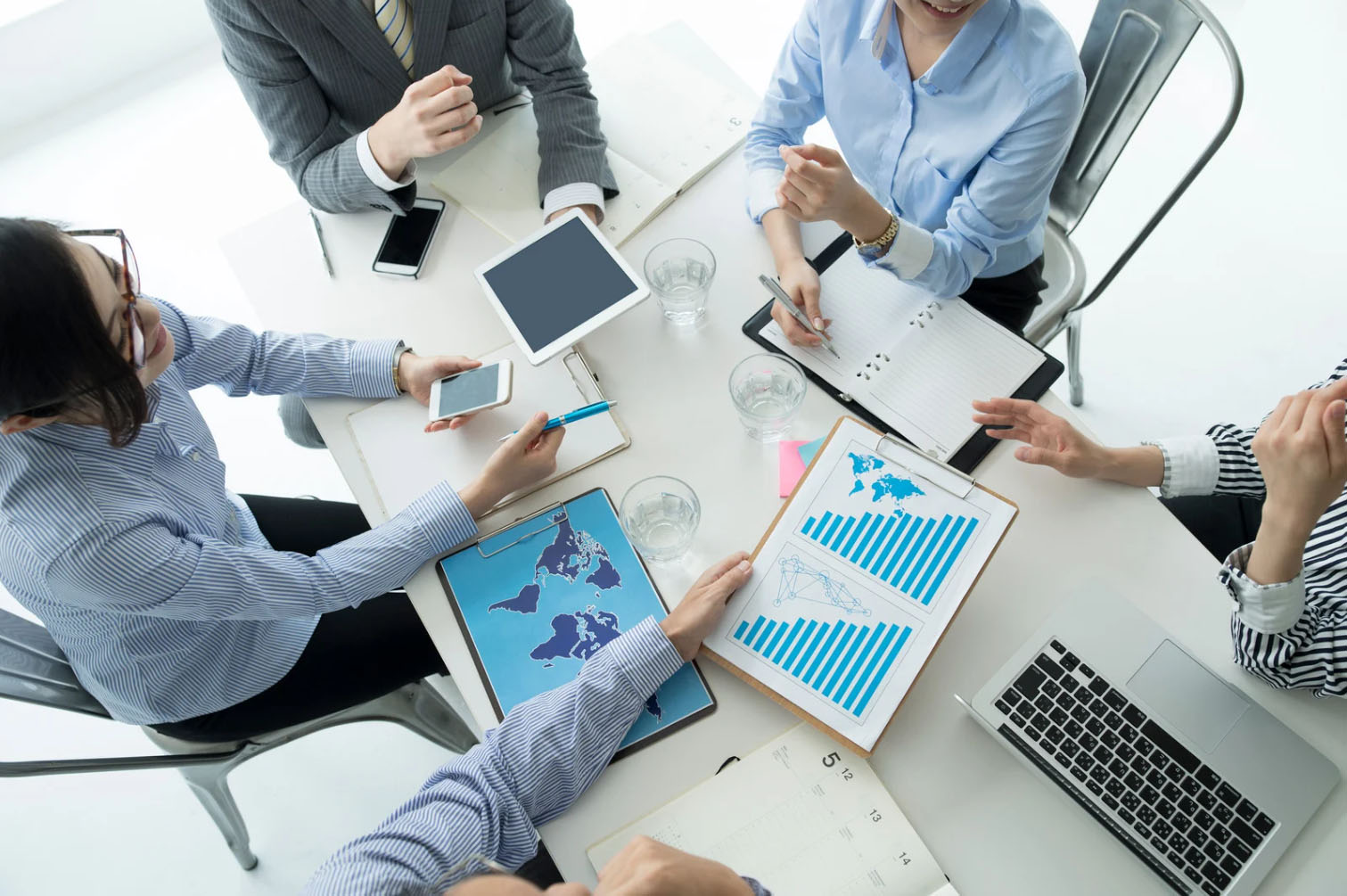 A group of professionals in a business meeting, discussing documents with charts, maps, and using smartphones and laptops.