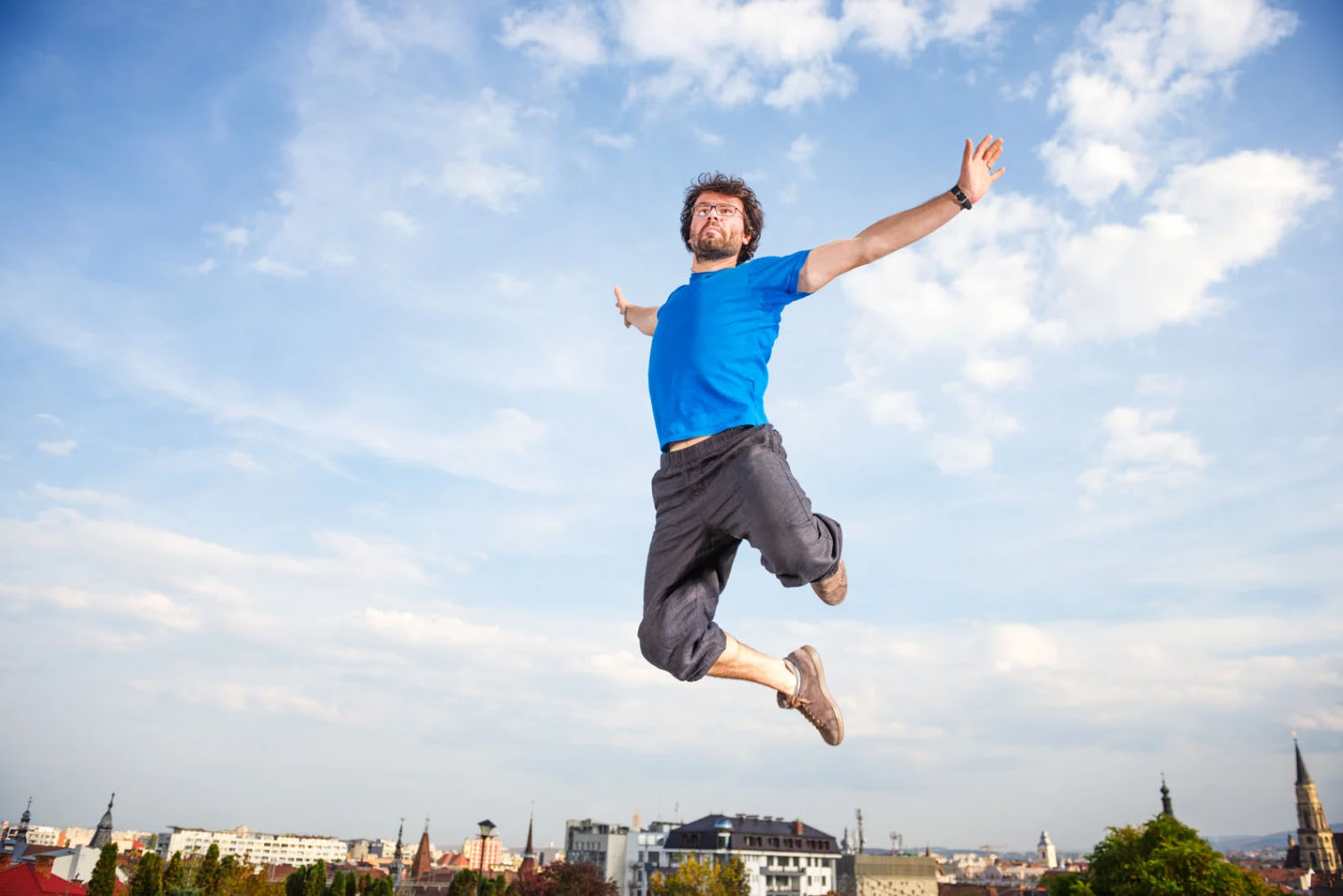 A man wearing a blue shirt and gray pants jumps energetically in the air against a bright blue sky, symbolizing vitality, freedom, and renewed energy.