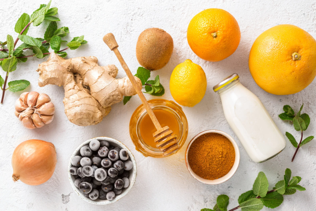 Flat lay of immune-boosting foods including ginger, garlic, onion, blueberries, citrus fruits, kiwi, honey, turmeric powder, milk, and fresh mint leaves on a white background.