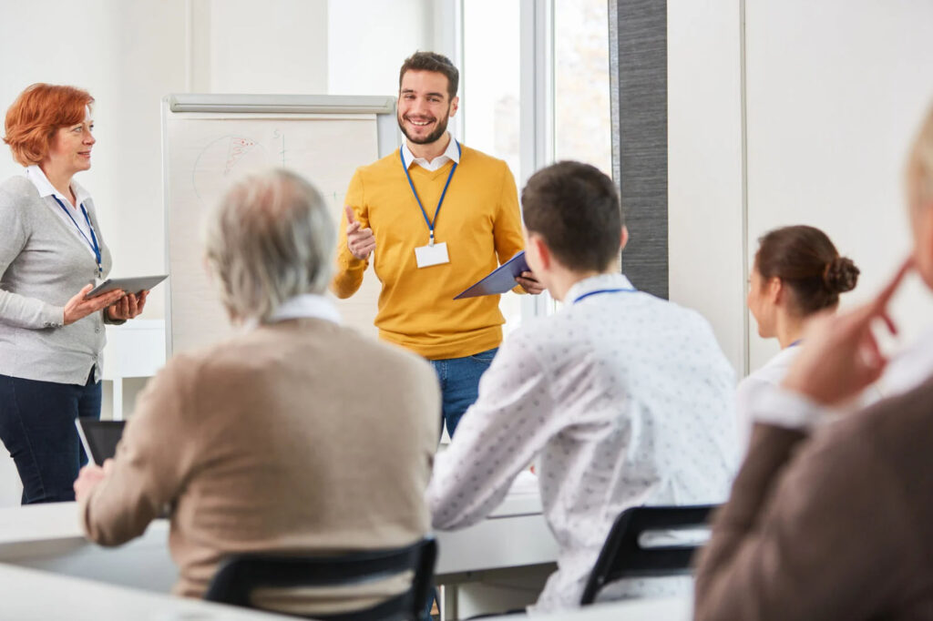 A group of professionals attending a seminar. The speaker, a man in a yellow sweater and a name tag, is presenting to the audience. A woman in a white shirt is standing beside him, holding a tablet. The audience, seated at a table, listens attentively.