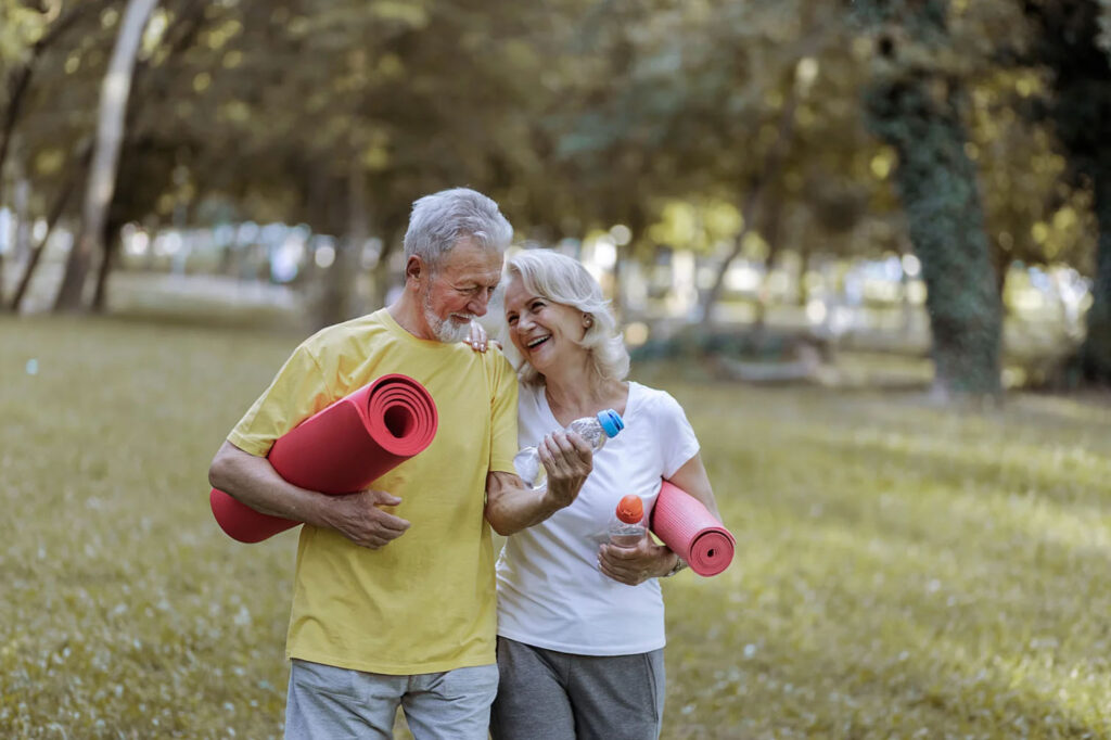 Smiling senior couple holding yoga mats and water bottles while walking together in a park, enjoying outdoor exercise and wellness.