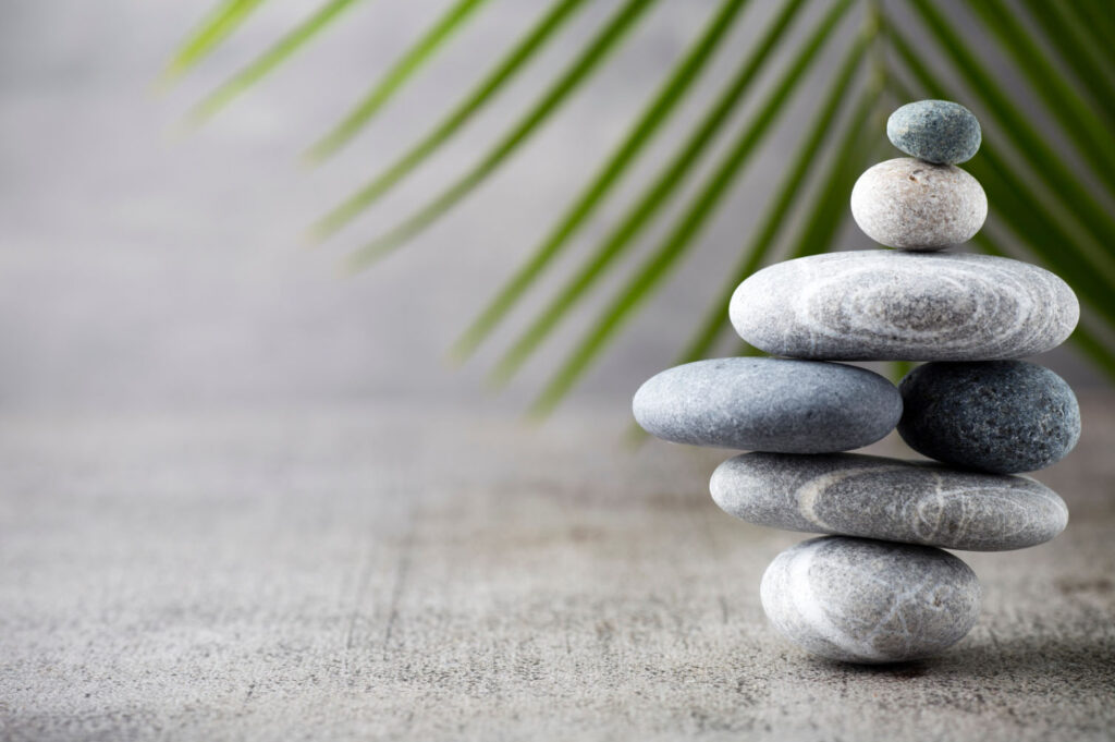 A serene image of stacked stones arranged in a balanced formation, with a blurred green palm leaf in the background.