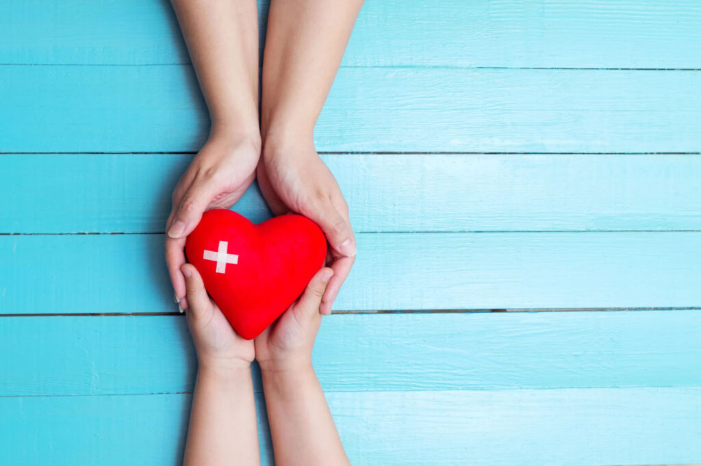 Two hands gently hold a red heart-shaped object with a white medical cross on it, symbolizing care and health, set against a light blue wooden background.